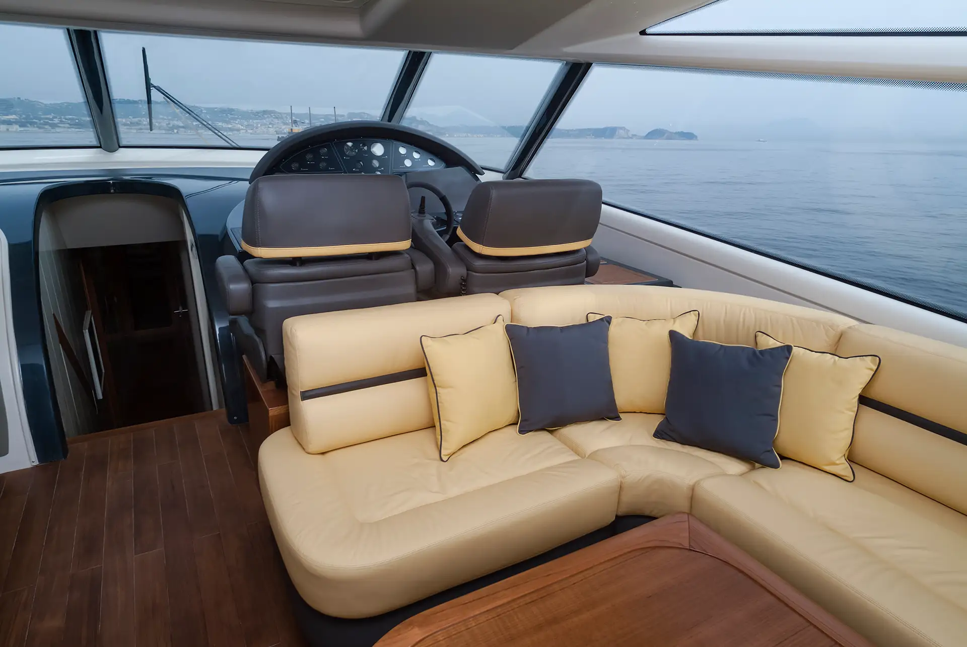 Yacht-Salon mit U-Sitzgruppe in beigem Leder und Holztisch; große Fensterfront mit Blick nach draußen und warmes, hochwertiges Interieur